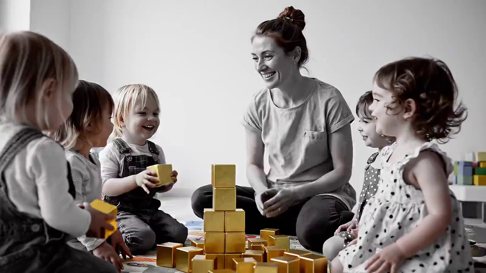 Een lachende begeleider zit op de vloer tussen een groepje peuters die met gouden bouwblokken spelen