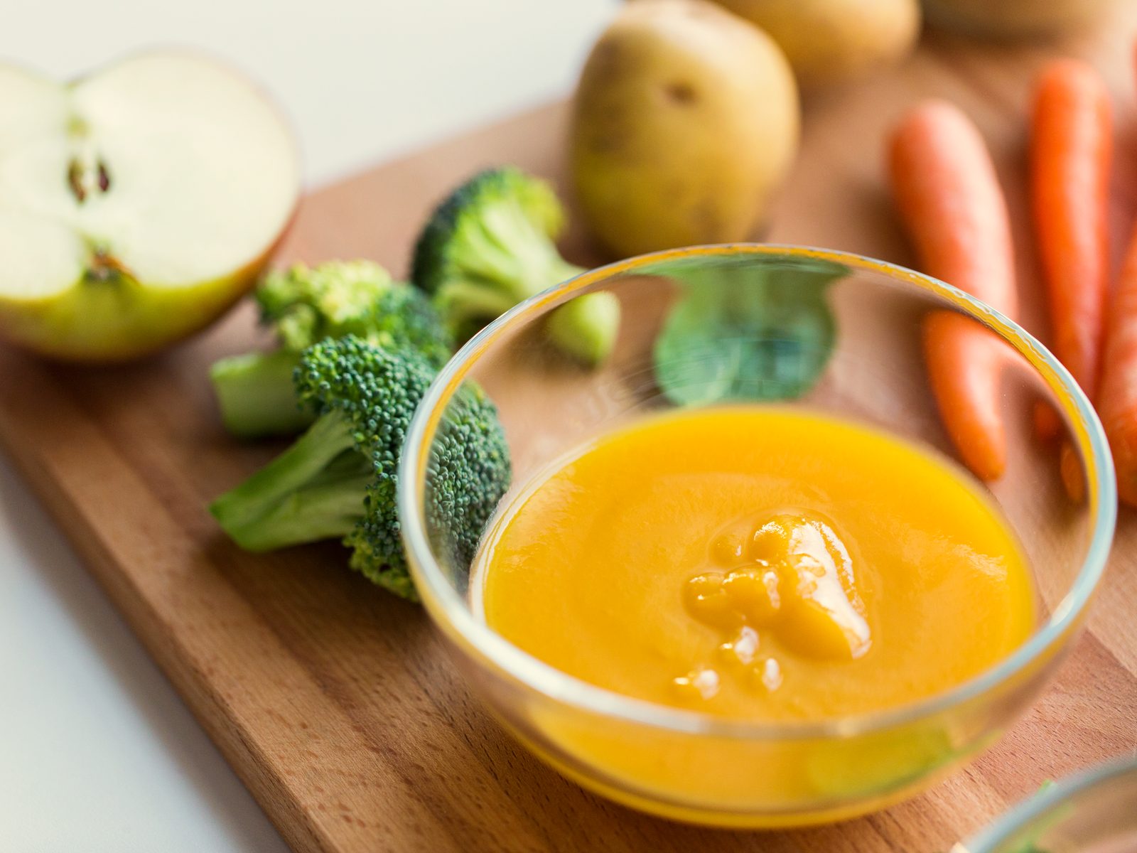 Kommetje groentepuree tussen broccoli, wortelen en een halve appel